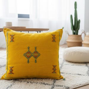 Peut inclure: Un coussin décoratif carré jaune avec des motifs géométriques brodés en rouge, vert et bleu. Le coussin est posé sur un tapis à motifs blancs et gris, avec un cactus et des paniers tressés en arrière-plan.