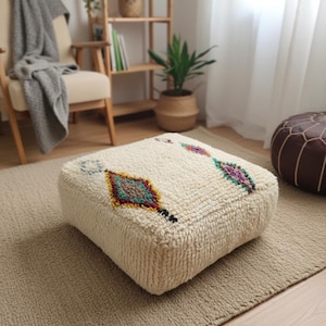 May include: A square, off-white Moroccan pouf with colorful geometric designs. The pouf is made of a textured, woven material and features diamond and rectangular patterns in shades of red, green, yellow, and purple. It sits on a neutral-colored rug.