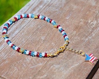 May include: A beaded bracelet with red, white, and blue beads, and gold star-shaped accents. A gold chain and clasp connect to a star charm with a red, white, and blue flag design. The bracelet is on a wooden surface.