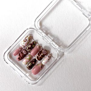 Könnte beinhalten: Ein Set von aufklebbaren Nägeln in einer transparenten Kunststoffbox. Die Nägel weisen verschiedene Designs auf, darunter braune Affen, rosa und weiße Punkte sowie rosafarbene Spitzen mit braunen Akzenten. Die Nägel sind oval geformt.