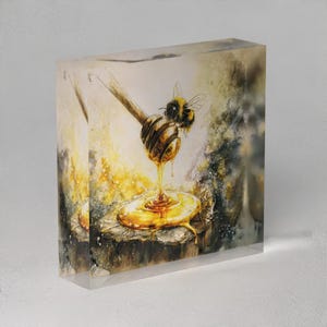 May include: A clear acrylic block featuring a watercolor painting of a bee hovering near a wooden honey dipper dripping honey onto a wooden surface. The artwork has warm yellow and brown tones.