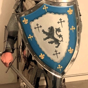 Könnte beinhalten: Ein mittelalterlicher Ritterschild mit blauem Hintergrund und einem silbernen Löwen mit schwarzem Schwanz und Mähne. Der Schild ist mit goldenen Lilien und silbernen Kreuzen verziert.
