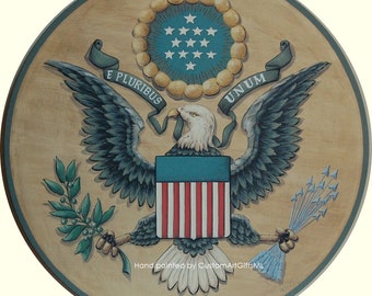 May include: A hand-painted wooden round sign featuring the Great Seal of the United States. The seal depicts a bald eagle with outstretched wings holding an olive branch in its right talon and arrows in its left talon. The eagle is perched on a shield with 13 red and white stripes and a blue field with 13 white stars. The seal is surrounded by a wreath of olive branches and a ribbon with the words "E Pluribus Unum".