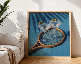 May include: A framed print depicting two martini glasses with olive skewers, placed on a vintage tennis racket. The artwork has a blue background with palm frond shadows. The frame is made of light-coloured wood.