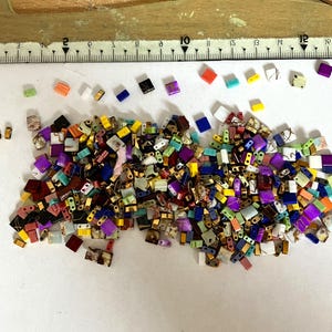 Peut inclure: Un assortiment de petites perles rectangulaires de différentes couleurs, dont le violet, le bleu, le jaune et le blanc, avec des accents dorés. Une règle est visible en arrière-plan, indiquant la taille des perles. Les perles sont éparpillées sur une surface blanche.