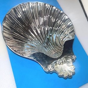 May include: A polished silver-toned metal dish shaped like a large scallop shell. The surface features deep, radiating ridges and a smooth, reflective finish. A smaller, decorative shell-shaped accent is attached at the base of the main bowl.