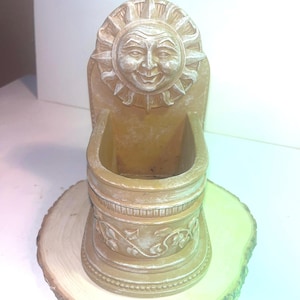 May include: A tan-colored wall-mounted planter featuring a sculpted sun face at the top and a vine pattern embossed around the base. The object has a semi-circular opening for holding items and a weathered finish with lighter highlights on the edges.