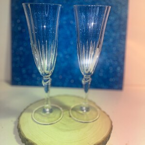May include: Two clear champagne flutes with vertical etched lines, resting on a round wooden slice. The flutes have a twisted stem and sit in front of a blue background with glitter.