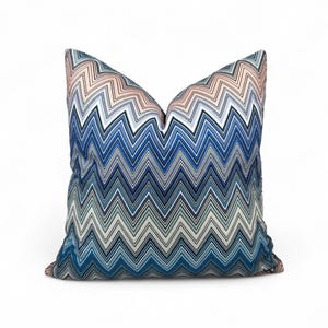 May include: A square cushion featuring a chevron pattern in shades of blue, white, and peach. The zig-zag design creates a modern look. The cushion is designed for home decor and has a soft, textured appearance.