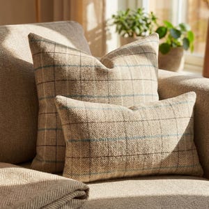 May include: Two beige plaid cushions with blue and brown accents sit on a beige sofa. The cushions are rectangular and square. A folded beige blanket is visible in the foreground. The scene is lit by natural light.