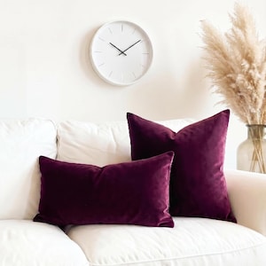 May include: Two deep purple velvet throw pillows, one square and one rectangular, resting on a white sofa. The pillows feature a soft, plush texture and a rich, dark plum color that contrasts with the bright white upholstery of the couch.