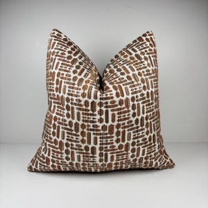 May include: A decorative throw pillow with a brown and white geometric pattern. The pillow has a textured appearance and a square shape. The pattern consists of repeating rectangular and oval shapes in varying shades of brown against a white background.