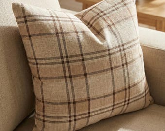 May include: A square decorative pillow with a plaid pattern in shades of beige and brown. The pillow is placed on a beige sofa. The pillow has a soft, textured appearance.