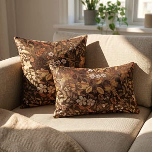 May include: Two decorative throw pillows with a floral pattern in shades of brown, beige, and white. The pillows are rectangular and square, resting on a neutral-colored sofa. The floral design includes leaves and flowers.