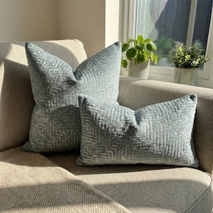 Puede incluir: Dos cojines decorativos verde azulado con un patr&oacute;n geom&eacute;trico sobre un sof&aacute; beige. Los cojines tienen una apariencia texturizada y est&aacute;n colocados frente a una ventana con plantas.