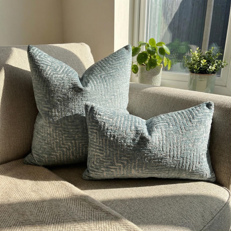 Puede incluir: Dos cojines decorativos verde azulado con un patr&oacute;n geom&eacute;trico sobre un sof&aacute; beige. Los cojines tienen una apariencia texturizada y est&aacute;n colocados frente a una ventana con plantas.