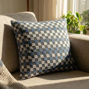May include: Square throw pillow with a checkered pattern in shades of gray, blue, and white. The pillow is placed on a neutral-toned sofa, with sunlight illuminating the fabric. The pillow adds a decorative element to the living room.
