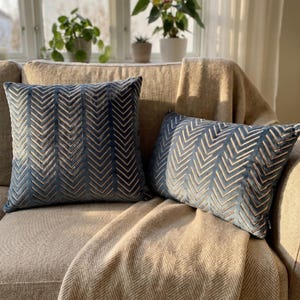 Peut inclure: Deux coussins en velours bleu ornés d'un motif chevron doré en relief. L'un est carré et l'autre rectangulaire. Les deux coussins présentent un motif en zigzag vertical cohérent sur leurs surfaces douces et texturées.