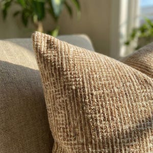 May include: Close-up of a textured, beige pillow with a woven pattern. The pillow has a pointed corner and is resting on a light-colored surface. The fabric appears to be a blend of beige and off-white threads.