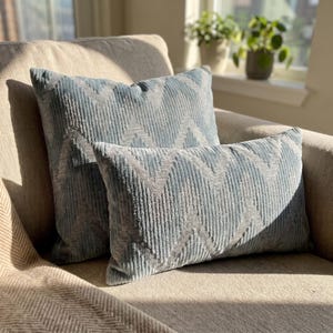 Puede incluir: Dos cojines decorativos de color azul claro con una superficie texturizada y acanalada. Los cojines presentan un patrón repetitivo de chevrón o zigzag en un tono de azul más claro. Un cojín es cuadrado y el otro es de forma rectangular.