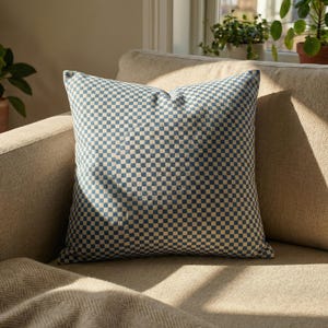 May include: A square cushion featuring a blue and beige chequered design. The cushion is positioned on a beige sofa, with a matching blanket. The pattern is a classic, geometric design, suitable for home decor.
