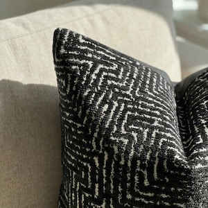 Op de afbeelding: Close-up van een decoratief kussen met een zwart-wit geometrisch patroon. Het kussen heeft een textuuroppervlak en is geplaatst op een neutraal gekleurde bank. Het ontwerp heeft een zich herhalend doolhofachtig motief.