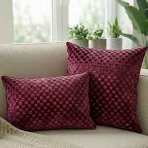 May include: Two burgundy throw pillows with a checkered pattern rest on a beige sofa. The pillows have a soft, velvety texture and a square design. The pillows are different sizes, one rectangular and one square.