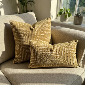 Könnte beinhalten: Drei dekorative Kissen in Gold- und Cremetönen mit einem geometrischen Muster liegen auf einem Sofa. Die Kissen haben eine strukturierte Oberfläche. Das Arrangement umfasst ein rechteckiges und zwei quadratische Kissen.
