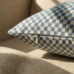 May include: A close-up shot of a cushion with a blue and beige chequered pattern. The cushion has a zip closure and is on a beige surface. The pattern is a repeating grid of blue and beige squares.