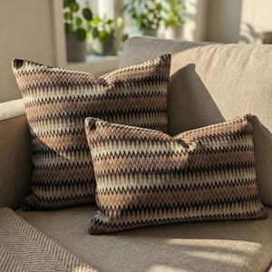 Może przedstawiać: Dwie dekoracyjne poduszki z brązowo-beżowym zygzakowatym wzorem spoczywają na beżowej sofie. Poduszki mają teksturowany wygląd i są prostokątne. Dodają ciepła i stylu do salonu.