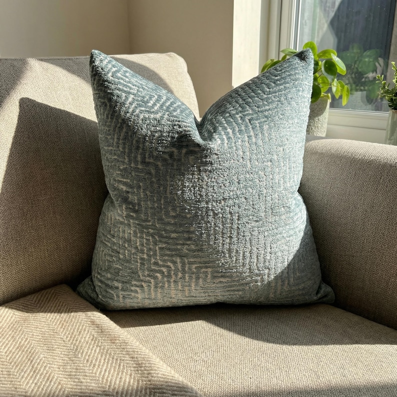 Puede incluir: Un coj&iacute;n cuadrado azul claro con un patr&oacute;n geom&eacute;trico. El coj&iacute;n est&aacute; colocado sobre un sof&aacute; beige, con la luz del sol resaltando la tela texturizada. El dise&ntilde;o del coj&iacute;n a&ntilde;ade un toque de elegancia a la decoraci&oacute;n de la habitaci&oacute;n.