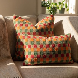May include: Two rectangular throw pillows with a vibrant checkerboard design in red, orange, green, and tan. The cushions are placed on a neutral-toned sofa. A folded blanket is visible on the left side of the image.