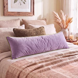 Pode incluir: Uma almofada retangular longa de veludo lavanda com uma borda fina verde. A almofada está posicionada horizontalmente sobre uma cama com roupa de cama em tons neutros, incluindo uma almofada castanha texturizada e uma manta com padrão.