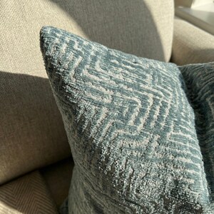 Puede incluir: Primer plano de un coj&iacute;n decorativo azul con un patr&oacute;n geom&eacute;trico. El coj&iacute;n tiene una superficie texturizada y un dise&ntilde;o en zigzag azul claro y blanco. El coj&iacute;n est&aacute; sobre un sof&aacute; beige.