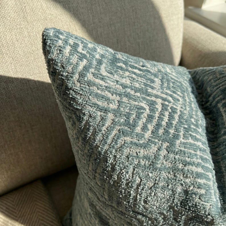 Puede incluir: Primer plano de un coj&iacute;n decorativo azul con un patr&oacute;n geom&eacute;trico. El coj&iacute;n tiene una superficie texturizada y un dise&ntilde;o en zigzag azul claro y blanco. El coj&iacute;n est&aacute; sobre un sof&aacute; beige.