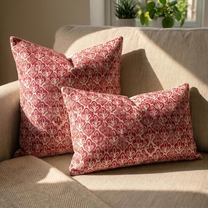 May include: Two throw pillows featuring a repeating red and cream botanical leaf pattern. One pillow is square and the other is rectangular. Both cushions have a textured surface and are displayed together on a neutral-colored fabric sofa.