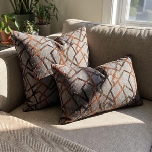 Puede incluir: Dos cojines decorativos de tela gris texturizada con un patrón geométrico de líneas entrelazadas en tonos cobre y marrón oscuro. El conjunto incluye un cojín cuadrado y uno rectangular, ambos con el mismo diseño tejido.
