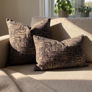 Peut inclure: Deux coussins décoratifs présentant un motif de grille abstrait marron foncé et beige. L'un est carré et l'autre est rectangulaire. Les deux coussins ont une surface en tissu texturé et sont posés sur un canapé beige clair à chevrons.