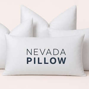 May include: Several white pillows of varying shapes and sizes are arranged against a light background. The central pillow displays the text "NEVADA PILLOW" in a dark blue font, indicating a home decor item.