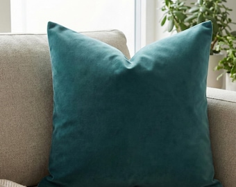 Puede incluir: Un cojín cuadrado de color verde azulado con una textura suave y aterciopelada descansa sobre un sofá de tonos neutros. El color intenso del cojín contrasta con la tela más clara del sofá, creando un acento visualmente atractivo. El cojín es un elemento decorativo para la decoración del hogar.