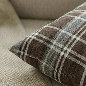 Pode incluir: Close-up de uma almofada xadrez marrom, cinza e branca. A almofada tem um padrão xadrez clássico com linhas que se cruzam. A almofada é feita de um tecido texturizado e é colocada em uma superfície de cor neutra.