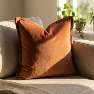 Puede incluir: Un cojín cuadrado de terciopelo naranja quemado con un borde marrón oscuro. El cojín está colocado en un sofá gris claro, con la luz del sol iluminando su superficie texturizada. Acento decorativo para el hogar.