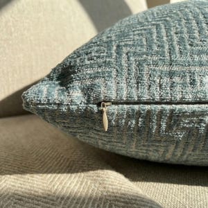 Puede incluir: Primer plano de un coj&iacute;n decorativo azul verdoso con un patr&oacute;n geom&eacute;trico texturizado. El coj&iacute;n tiene un cierre de cremallera con un tirador plateado. El coj&iacute;n descansa sobre una superficie de tela clara.