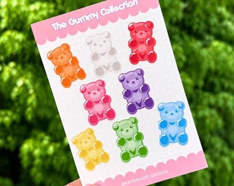 May include: A pink card with illustrations of gummy bears in various colours. The bears are orange, white, red, pink, purple, blue, yellow, and green. The text "The Gummy Collection" is at the top, and "@PAPERHARTSDESIGN" is at the bottom.