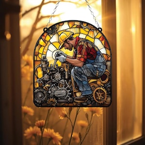 May include: Stained glass window art depicting a mechanic working on machinery. The artwork features a man in a yellow hard hat and overalls, with a red and black plaid shirt. The background is a warm yellow and orange, with gears and cogs.