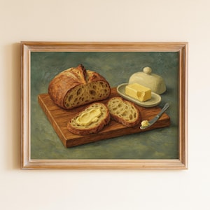 Op de afbeelding: Een ingelijst schilderij toont een brood, gesneden brood, boter en een botervloot. Het brood ligt op een houten snijplank. Er is ook een mes met boter aanwezig. De achtergrond is een gedempt groen.