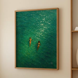 May include: A framed painting depicts two figures floating in turquoise water. The figures are wearing swimwear. The artwork is in a wooden frame and is displayed on a white wall.