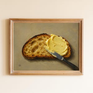 Puede incluir: Pintura enmarcada de una rebanada de pan tostado con mantequilla untada. El pan tiene una corteza oscura y un interior texturizado. La mantequilla es de un amarillo brillante, y se utiliza un cuchillo con mango negro para untarla.