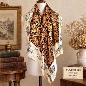May include: A square silk scarf featuring a central brown and black leopard print pattern surrounded by a cream-coloured border decorated with black and white illustrations of leopards. The scarf measures 86.36 by 86.36 cm as stated on the sign.