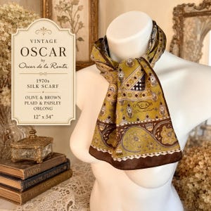 May include: An oblong silk scarf by Oscar de la Renta featuring an olive green and brown color palette with a detailed paisley and plaid pattern. The scarf displays the Oscar de la Renta brand name printed on the fabric and measures 12 by 54 inches.